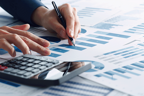 Detail image of a spreadsheet on a table along with a calculator and a person’s hand making notes on the spreadsheet.